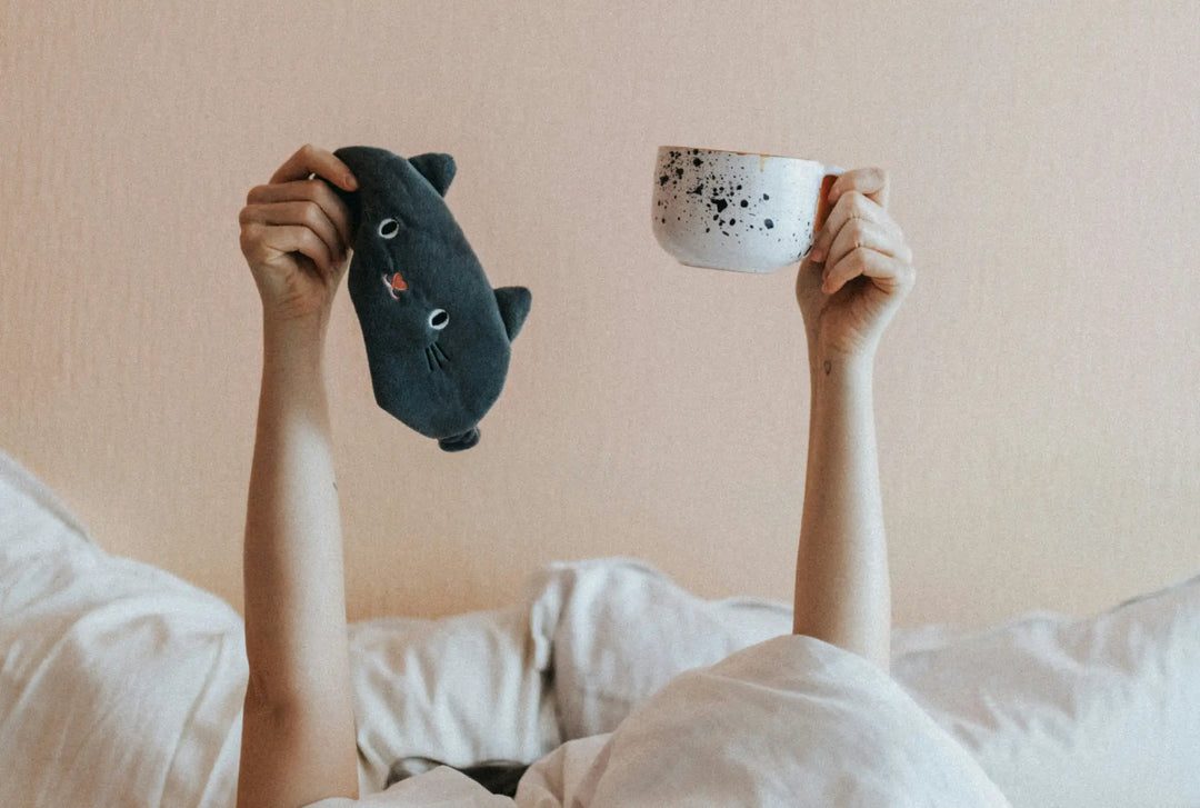 A person holding a mug and a sleep mask above their head while lying in bed