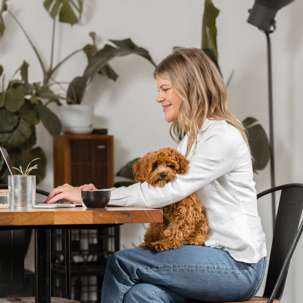 A woman sitting at a table in a cafe, with a dog on her lap, holding a cup of Earthrise and smiling