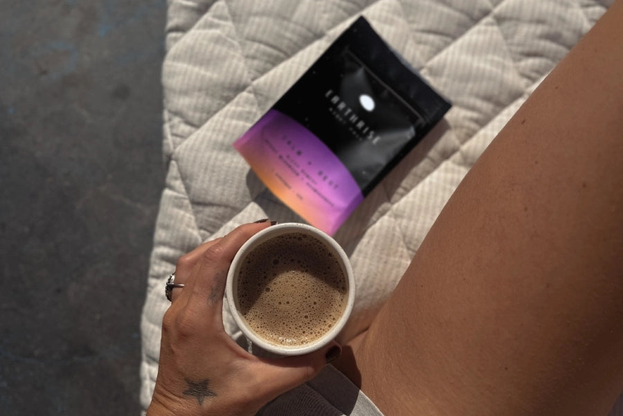 A picture of a hand holding a cup of coffee next to a thight with a blurred packet of Earthrise coffee in the background on a quilted bed spread. 